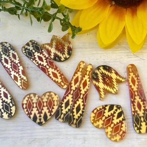May include: A collection of hair clips in various shapes including hearts, tulips, and elongated teardrops. Each piece features a repeating geometric pattern in shades of dark red, black, and cream, finished with a smooth, glossy surface texture.