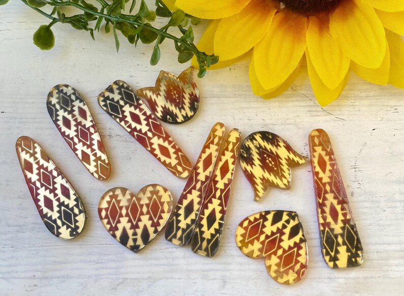 May include: A collection of hair clips in various shapes including hearts, tulips, and elongated teardrops. Each piece features a repeating geometric pattern in shades of dark red, black, and cream, finished with a smooth, glossy surface texture.