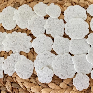 May include: A collection of white felt shapes with various designs, including flowers, strawberries, and leaves. The shapes are arranged on a woven wicker surface.
