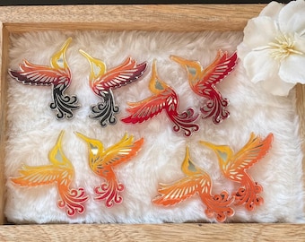 May include: A collection of hummingbird-shaped ornaments in shades of yellow, orange, and red, with intricate wing and tail details. The ornaments are displayed on a fluffy white surface within a wooden frame, with a white flower in the corner.