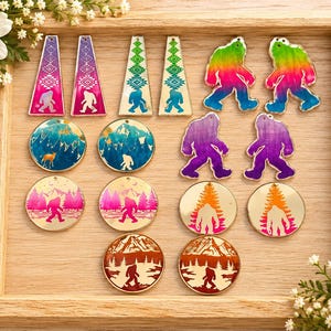 May include: A collection of assorted acrylic earring charms featuring various designs including Bigfoot silhouettes, mountain landscapes, and geometric patterns in vibrant colors like rainbow, purple, blue, pink, and orange, arranged on a wooden tray.