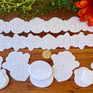 May include: A collection of white felt applique shapes featuring intricate floral and tulip line patterns. The set includes two long scalloped strips, several large floral motifs, and smaller circular pieces, all arranged with a small gold coin.