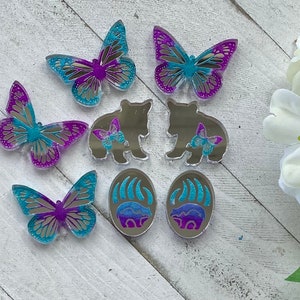 Op de afbeelding: Set van acht hars ornamenten met een berenpootafdruk, een beer met een vlinder en een vlinder. De ornamenten zijn in tinten blauw, paars en zilver.