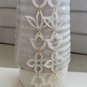 May include: A pair of long, white, dangling earrings with a floral design. The earrings are made of multiple layers of delicate, cut-out shapes, including tulips and flowers. The earrings are suspended from gold-colored hooks.