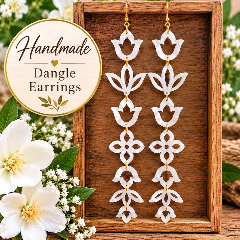 May include: A pair of long dangle earrings featuring white, pearlescent, cutout floral and geometric shapes linked together by gold-toned metal rings. The earrings hang from gold-toned hooks. A circular graphic with the text Handmade Dangle Earrings is visible in the upper left corner.
