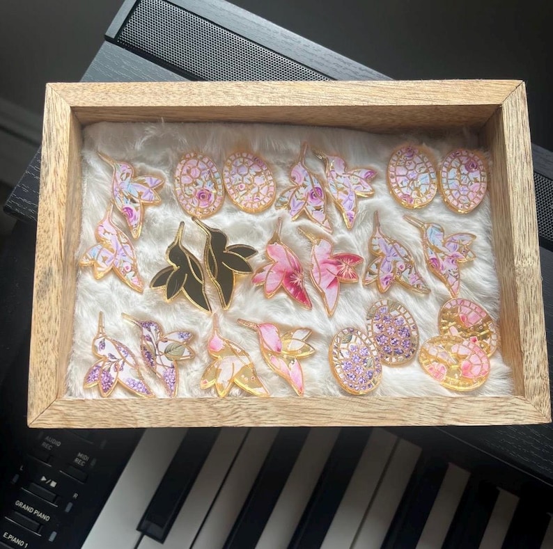 May include: A wooden display box filled with various hummingbird and floral-themed pins and earrings. The pins feature gold outlines and colorful enamel, including pink, purple, and black designs. The earrings have similar designs with a gold border.