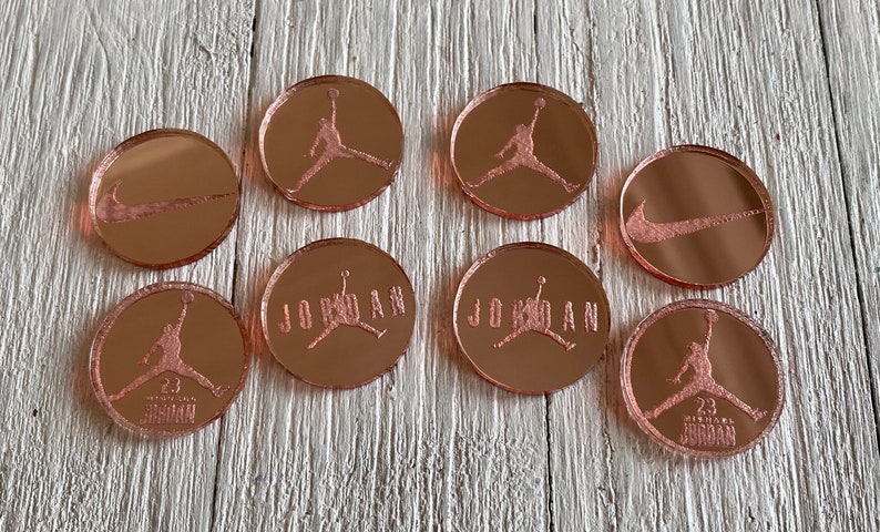 K&ouml;nnte beinhalten: Acht runde Spiegelkacheln in Ros&eacute;gold mit verschiedenen Designs. Die Designs umfassen den Nike-Swoosh, das Jordan-Logo und den Text "JORDAN" mit dem Jordan-Logo.
