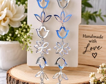 May include: A pair of long, silver-toned dangling earrings featuring a vertical arrangement of interconnected floral and leaf-shaped cutouts. The reflective, metallic pieces are linked together to create a cascading design that hangs from standard earring hooks.