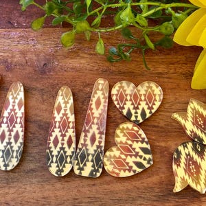 May include: A collection of flat, decorative acrylic shapes featuring a repeating geometric pattern in shades of cream, brown, and black. The set includes elongated teardrop pieces, two heart-shaped pieces, and two tulip-shaped pieces with smooth edges.