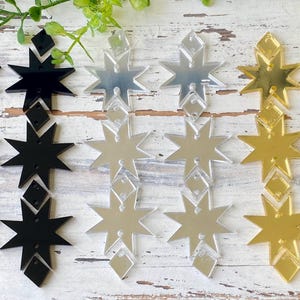 May include: A collection of star-shaped acrylic earring components arranged in three vertical pairs. The set includes two black, two silver mirrored, and two gold mirrored pairs, each featuring a multi-pointed star design with small diamond-shaped accents.