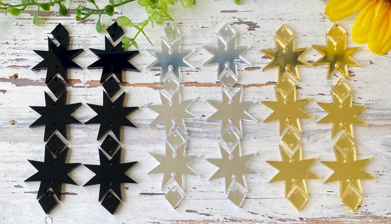 May include: A collection of star-shaped acrylic earring components arranged in three vertical pairs. The set includes two black, two silver mirrored, and two gold mirrored pairs, each featuring a multi-pointed star design with small diamond-shaped accents.