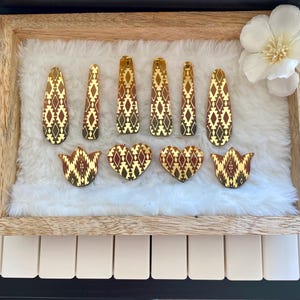 May include: A collection of ten hair clips featuring a reflective gold and dark brown geometric diamond pattern. The set includes six elongated teardrop-shaped clips, two heart-shaped clips, and two tulip-shaped clips, all arranged on a white surface.