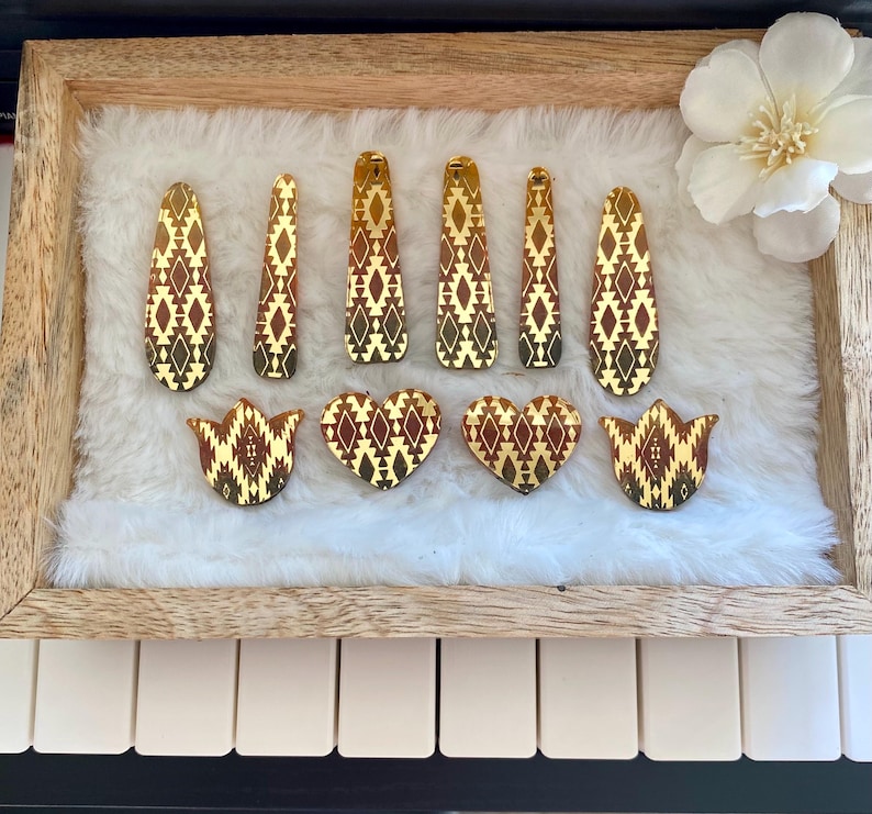 May include: A collection of ten hair clips featuring a reflective gold and dark brown geometric diamond pattern. The set includes six elongated teardrop-shaped clips, two heart-shaped clips, and two tulip-shaped clips, all arranged on a white surface.
