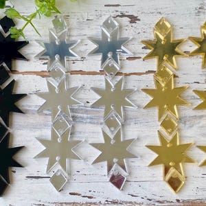 May include: A collection of star-shaped acrylic components arranged in three vertical columns. The set includes black, silver mirrored, and gold mirrored finishes. Each column features three eight-pointed stars connected by small diamond-shaped pieces.