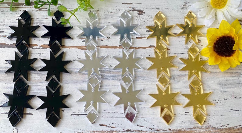 May include: A collection of star-shaped acrylic components arranged in three vertical columns. The set includes black, silver mirrored, and gold mirrored finishes. Each column features three eight-pointed stars connected by small diamond-shaped pieces.