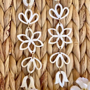 May include: A pair of long, dangling earrings featuring a series of interconnected, white, pearlescent, cutout floral and petal-shaped components linked by small gold-toned metal rings, creating a vertical, symmetrical, and intricate design.