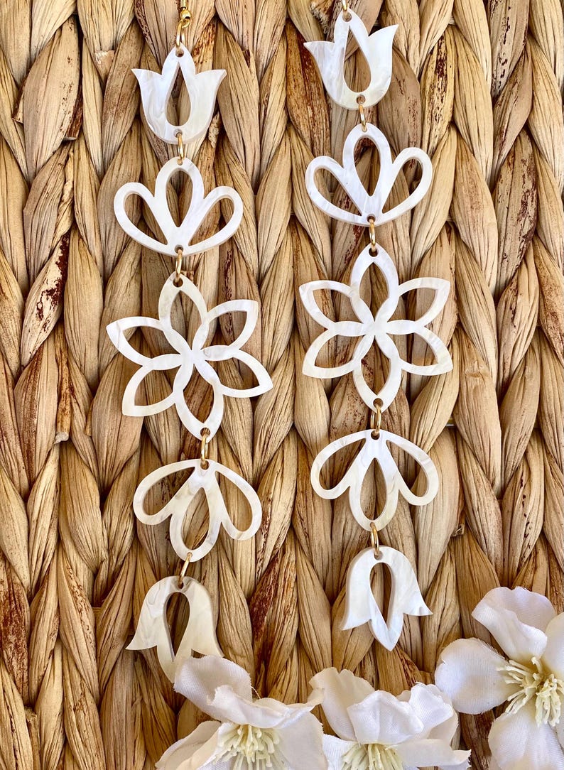 May include: A pair of long, dangling earrings featuring a series of interconnected, white, pearlescent, cutout floral and petal-shaped components linked by small gold-toned metal rings, creating a vertical, symmetrical, and intricate design.