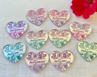 May include: Nine heart-shaped acrylic charms featuring a floral design and the word Indigenous printed in the center. The charms display a gradient color scheme transitioning from purple and pink to teal and green, arranged on a white surface.
