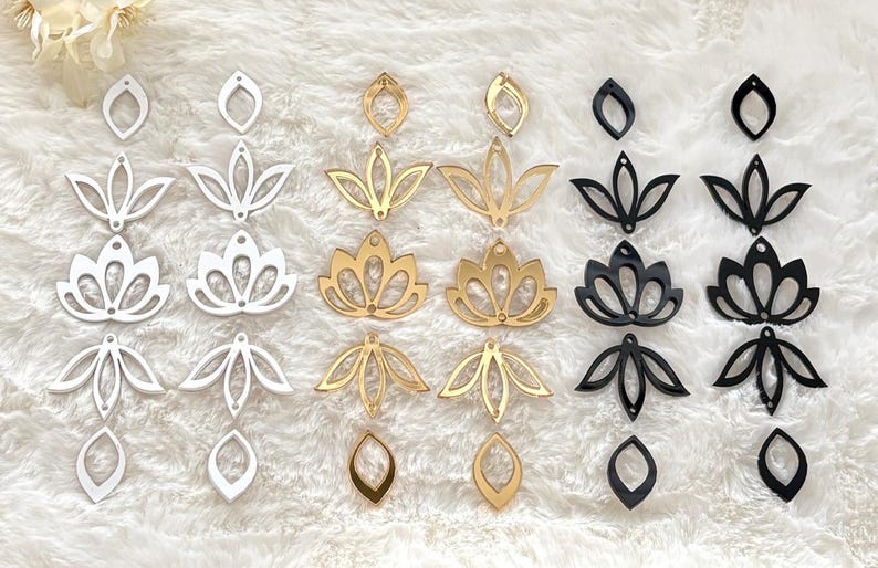 May include: A collection of laser-cut acrylic components in white, gold, and black. The set includes various lotus flower shapes, petal motifs, and teardrop-shaped connectors, each featuring small pre-drilled holes for assembly into jewelry pieces.