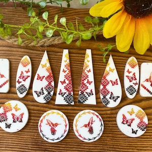 May include: A collection of white jewelry components featuring a gradient print of hummingbirds, butterflies, and geometric patterns in shades of yellow, orange, red, and black. The set includes various shapes such as teardrops, triangles, and circles.