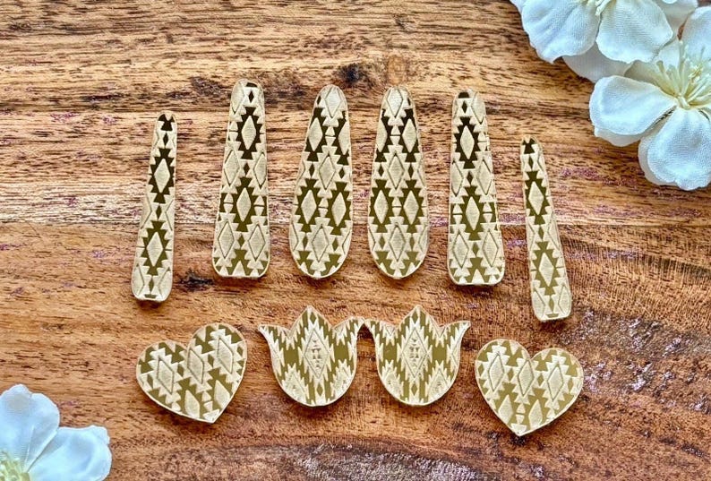 May include: A collection of ten flat, gold-toned decorative shapes featuring a repeating geometric diamond pattern. The set includes six elongated, tapered strips, two heart-shaped pieces, and two tulip-shaped pieces, all with a consistent metallic finish.