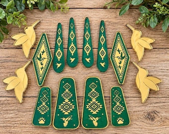 May include: A collection of jewelry components including four gold-toned mirrored hummingbird shapes, two diamond-shaped green pendants with gold hummingbird motifs, four long green teardrop pendants, and four trapezoidal green pendants with gold patterns.
