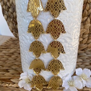 May include: A pair of long gold-toned dangle earrings featuring four stacked, petal-shaped segments per earring. Each segment has a reflective, metallic finish with a dark, intricate geometric diamond pattern etched into the surface.