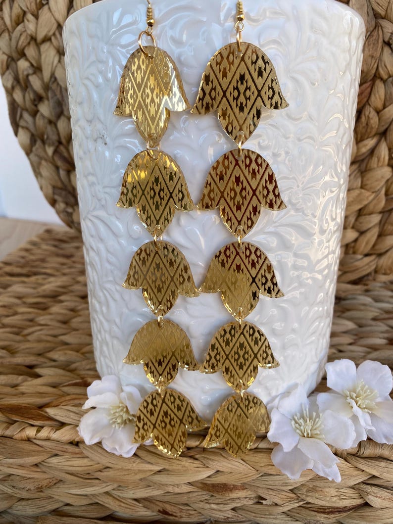 May include: A pair of long gold-toned dangle earrings featuring four stacked, petal-shaped segments per earring. Each segment has a reflective, metallic finish with a dark, intricate geometric diamond pattern etched into the surface.