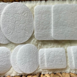 May include: An assortment of white, circular and square, fabric coasters with a floral design. The coasters are arranged on a white, fluffy surface, with a woven basket visible in the background.
