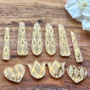 May include: A collection of ten gold-toned acrylic craft embellishments featuring a repeating geometric diamond pattern. The set includes six elongated teardrop shapes and four smaller pieces consisting of two hearts and two tulip-like floral shapes.