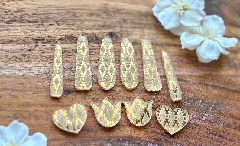 May include: A collection of ten gold-toned acrylic craft embellishments featuring a repeating geometric diamond pattern. The set includes six elongated teardrop shapes and four smaller pieces consisting of two hearts and two tulip-like floral shapes.