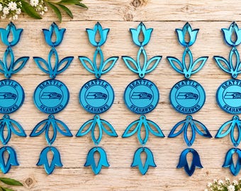 May include: Six identical blue acrylic ornaments arranged in two rows. Each ornament features a central circular disc with the Seattle Seahawks logo and text, flanked by symmetrical floral-shaped cutouts at the top and bottom of the design.