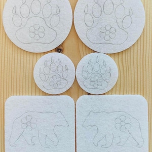 Puede incluir: Un conjunto de recortes de fieltro blanco con huellas de patas de oso y siluetas de osos con detalles florales. Los recortes, de formas circulares y cuadradas, están dispuestos sobre una superficie de madera clara.