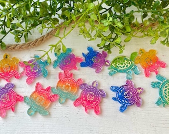 May include: A collection of twelve flat turtle-shaped charms featuring vibrant gradient colors including pink, orange, yellow, blue, and green. Each turtle has a detailed floral pattern on its shell and a small hole at the top for hanging.