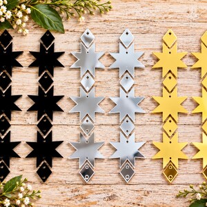 May include: Six vertical earring components arranged in three pairs of black, silver, and gold mirrored acrylic. Each piece features a repeating geometric design of three eight-pointed stars interspersed with small diamond-shaped connectors.