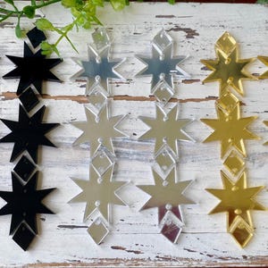 May include: A collection of six dangling earring components arranged in three pairs of black, silver, and gold star-shaped acrylic pieces. Each pair features two vertical star shapes connected by small diamond-shaped links with pre-drilled holes.