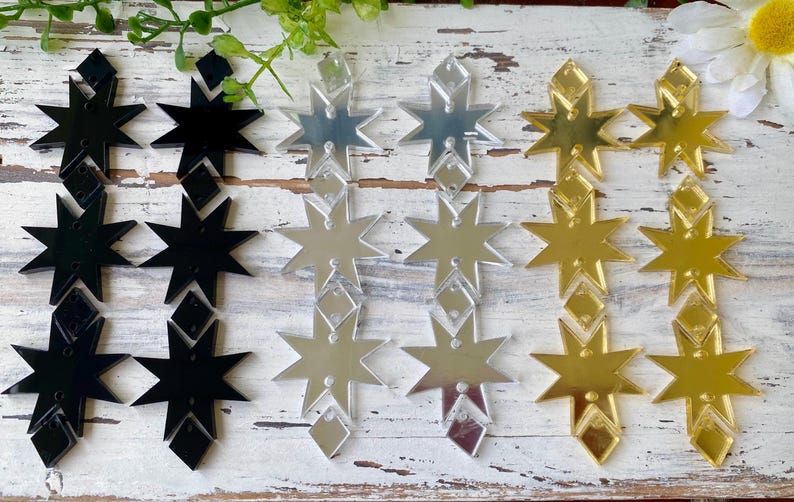 May include: A collection of six dangling earring components arranged in three pairs of black, silver, and gold star-shaped acrylic pieces. Each pair features two vertical star shapes connected by small diamond-shaped links with pre-drilled holes.