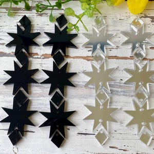 May include: A collection of eight-pointed star and diamond-shaped acrylic charms arranged in two columns. The left column features opaque black acrylic pieces, while the right column consists of reflective silver mirror acrylic pieces.