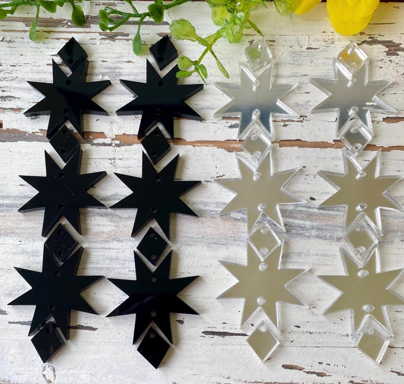 May include: A collection of eight-pointed star and diamond-shaped acrylic charms arranged in two columns. The left column features opaque black acrylic pieces, while the right column consists of reflective silver mirror acrylic pieces.