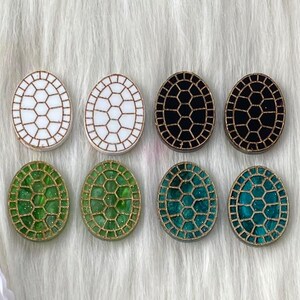 May include: Eight oval-shaped resin turtle shell earrings in white, black, and green with gold glitter accents.