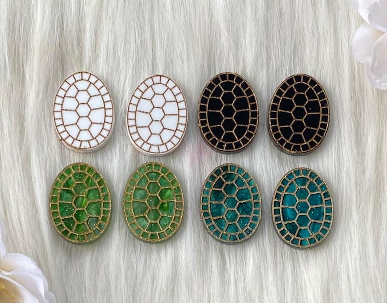 May include: Eight oval-shaped resin turtle shell earrings in white, black, and green with gold glitter accents.