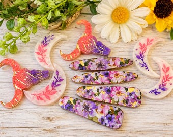 May include: A collection of resin accessories including two bull skull shapes with pink and purple gradients, two crescent moon shapes with pink and purple floral branch designs, and four hair clips featuring a vibrant purple and pink floral pattern.