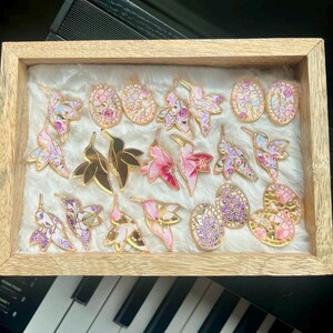 May include: A wooden framed display of decorative pins. The pins feature hummingbird, leaf, and oval designs with gold accents. The pins are in shades of pink, purple, and gold, set against a white, fluffy background.