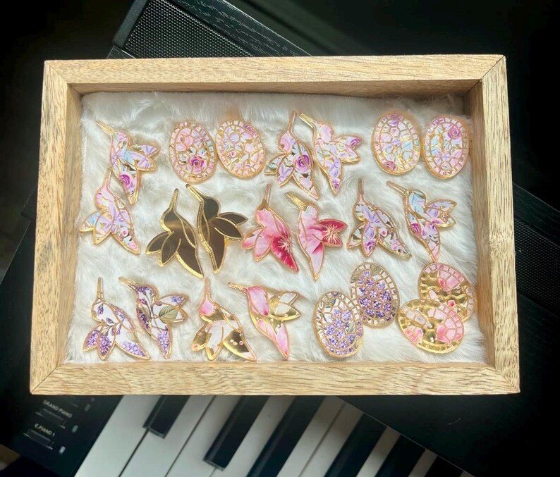 May include: A wooden framed display of decorative pins. The pins feature hummingbird, leaf, and oval designs with gold accents. The pins are in shades of pink, purple, and gold, set against a white, fluffy background.