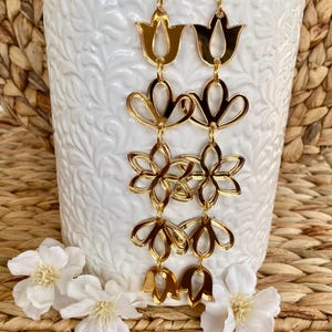 May include: A pair of long gold-toned dangle earrings featuring a vertical arrangement of five interconnected floral and tulip-shaped cutouts. The earrings have a reflective, metallic finish and are suspended from gold-toned earring hooks.