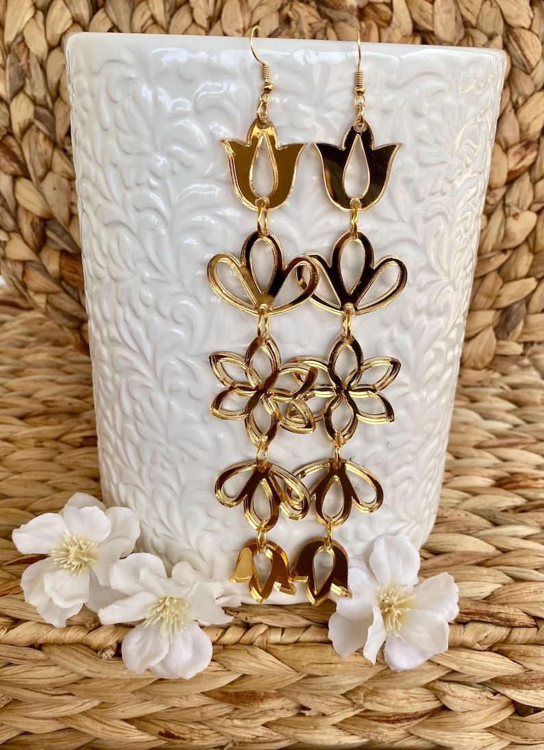 May include: A pair of long gold-toned dangle earrings featuring a vertical arrangement of five interconnected floral and tulip-shaped cutouts. The earrings have a reflective, metallic finish and are suspended from gold-toned earring hooks.