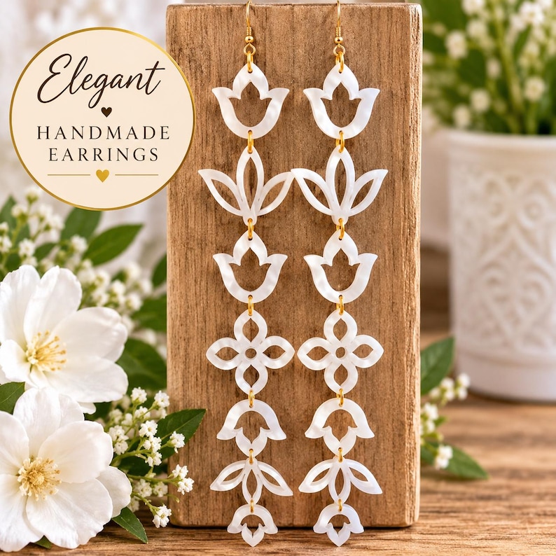 May include: Long drop earrings featuring a vertical arrangement of white, pearlescent, cutout floral and geometric shapes connected by gold-toned metal links. The earrings are displayed on a rectangular wooden block.