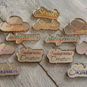 May include: A collection of 10 mirrored acrylic cloud-shaped ornaments with various phrases written on them, including "Indigenous Creature", "Native", and "Stoodis".