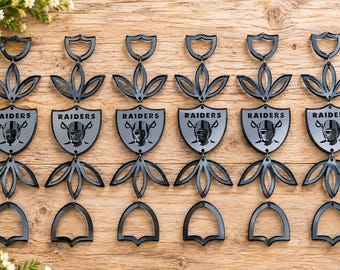 May include: Six identical black acrylic hanging ornaments arranged in a row. Each piece features a central shield-shaped emblem with the Raiders logo and text, flanked by decorative cut-out leaf patterns and topped with a small shield-shaped loop.