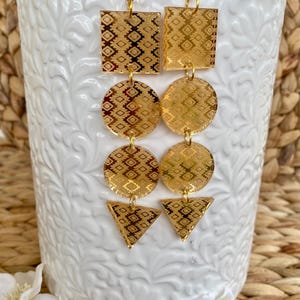 May include: A pair of gold-toned dangle earrings featuring a vertical arrangement of geometric shapes. Each earring consists of a square, two circles, and a triangle, all etched with a repeating diamond pattern and suspended from gold-toned hooks.